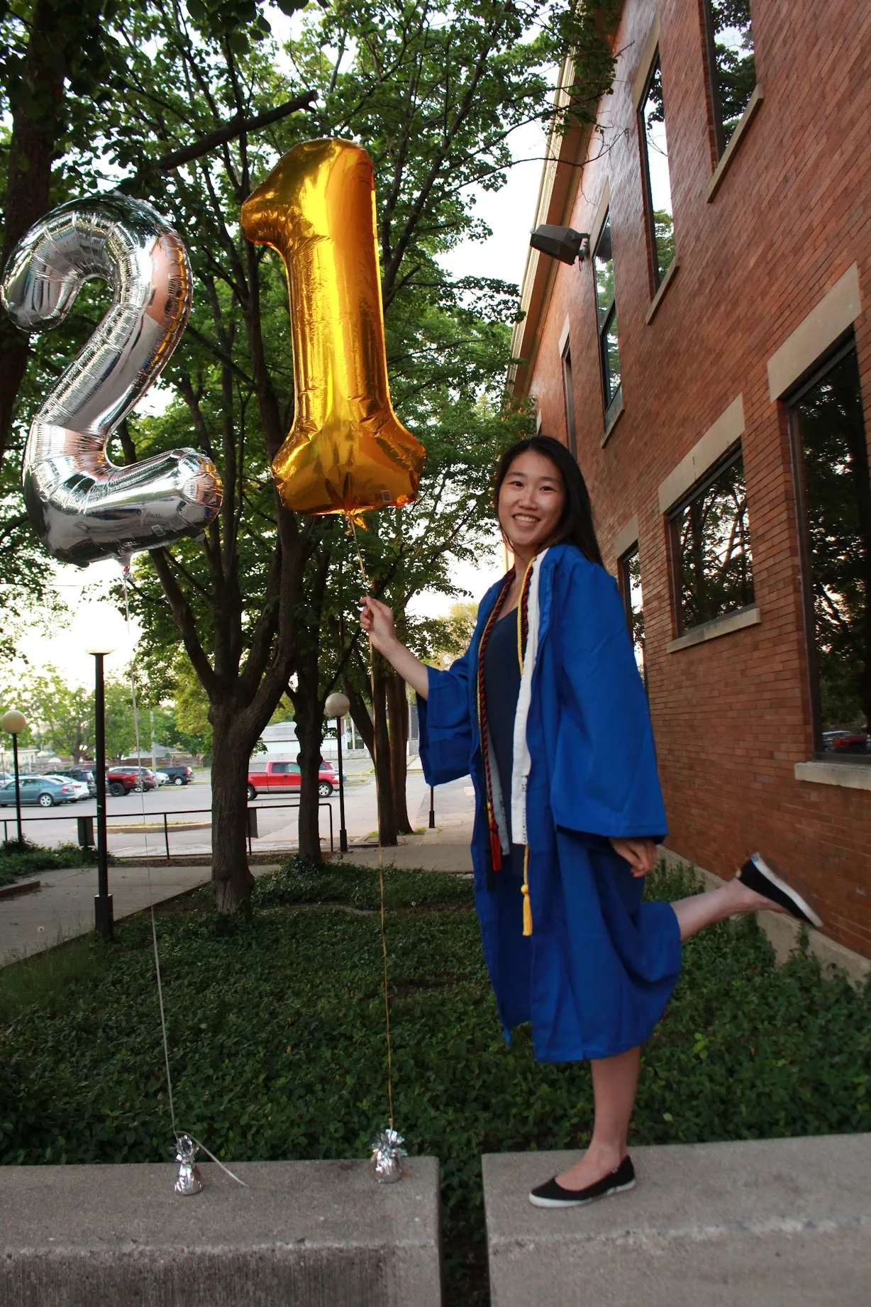 Xue holding silver '2' and gold '1' mylar balloons outside a brick campus building, wearing a blue graduation gown with honor cords and kicking one foot up.