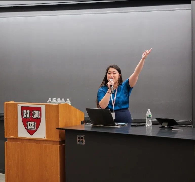 At a podium with the Harvard Veritas crest, mid-gesture during a talk