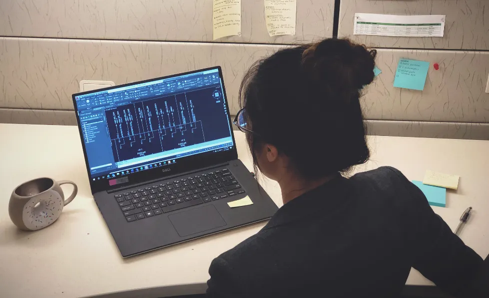 Xue seen from behind at a Herman Miller cubicle desk, working on a laptop showing an AutoCAD electrical schematic (not company info), with sticky notes pinned to the wall and a donut-print mug to the left.