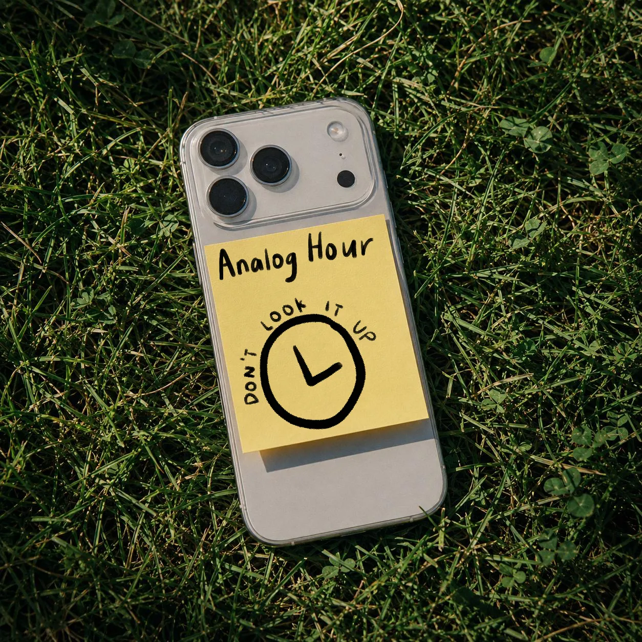An iPhone face-down on grass with a yellow sticky note on its back, hand-lettered "Analog Hour" above a drawn clock face circled by the words "don't look it up".