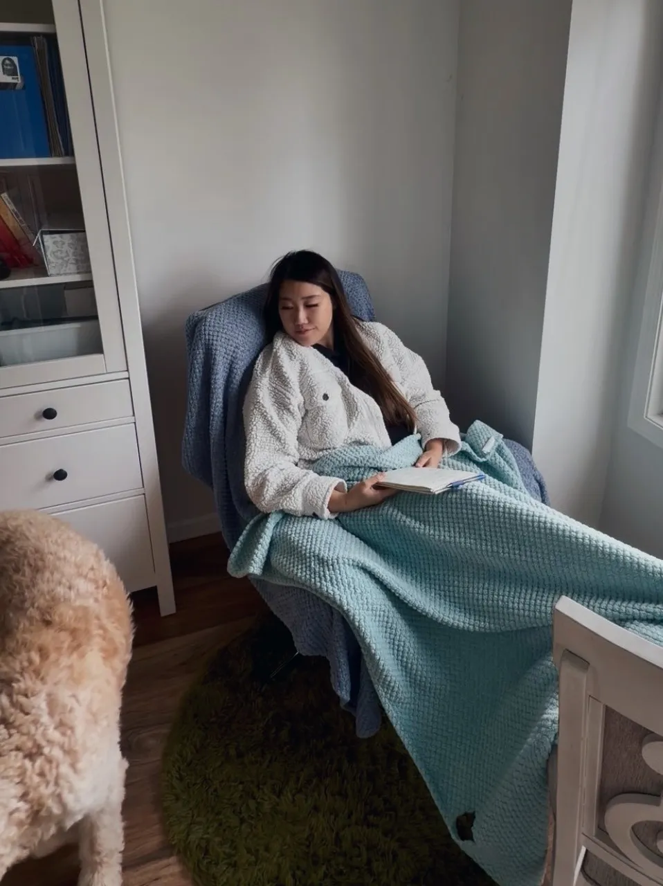 Xue reclined in a blue armchair by a window, tucked under a chunky cream-and-mint blanket with an open notebook and pen resting in my lap, eyes down and relaxed. A goldendoodle stands in the foreground.