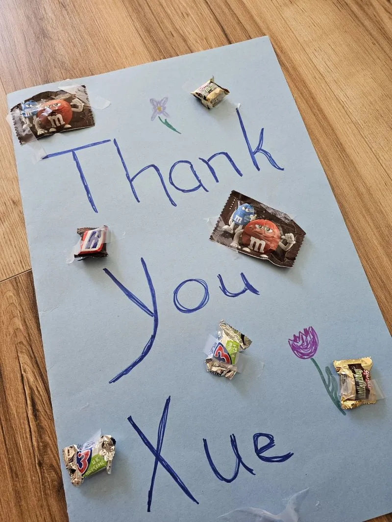 A handmade blue "Thank You Xue" sign decorated with taped-on candy wrappers and a drawn flower