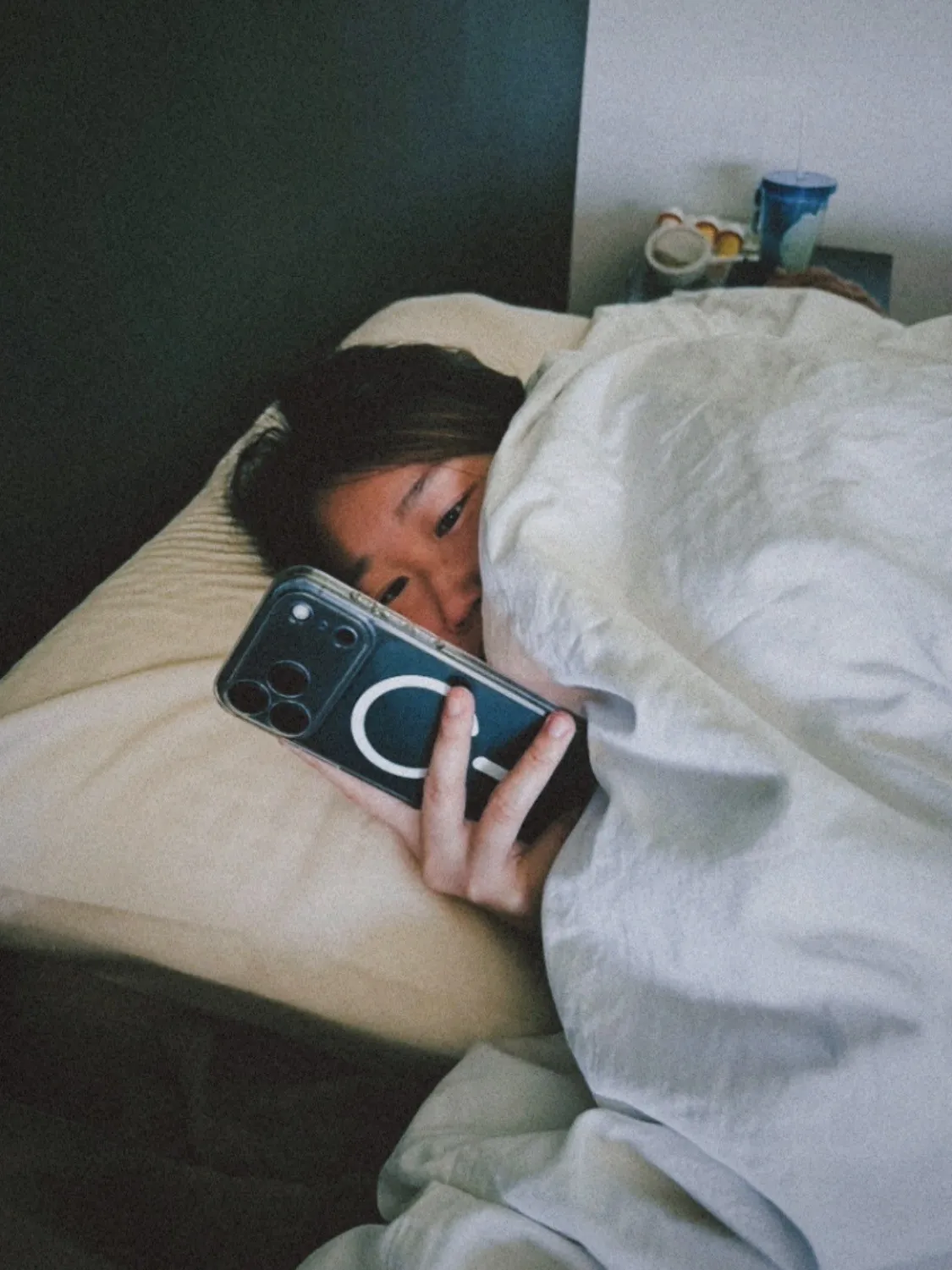 Xue lying in bed, half-tucked under a white duvet with cheek on the pillow, holding a phone close to my face in the dark.