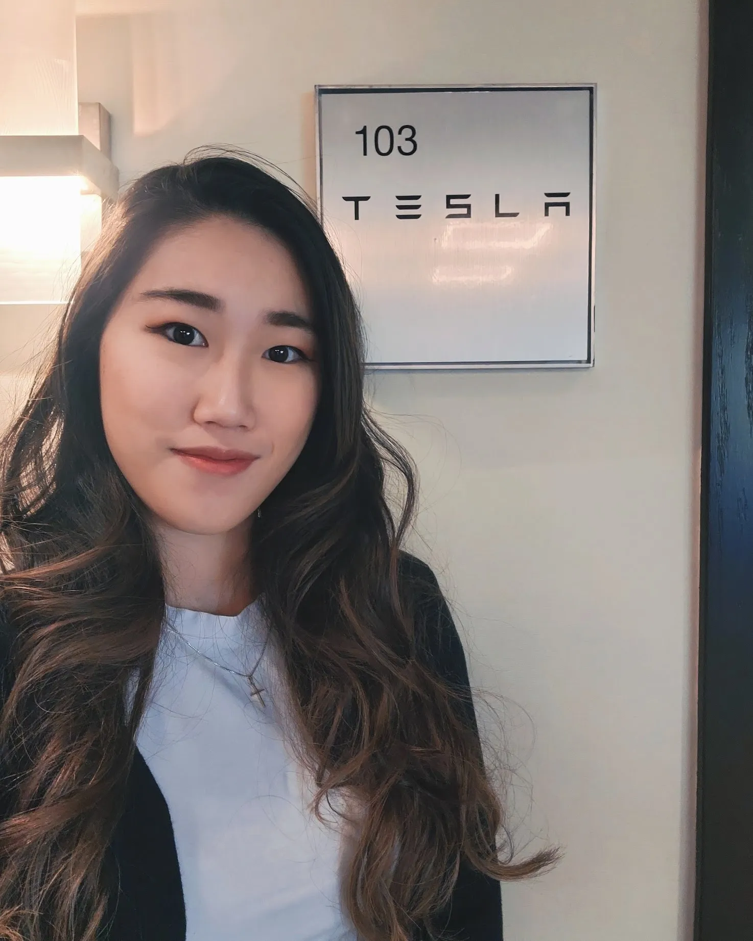 Xue smiling in front of a brushed-metal Tesla suite plaque numbered 103, mounted on a cream office wall.