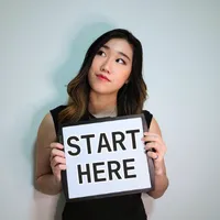 Photo of Xue holding a white card sign with the words START HERE in bold black capital letters, against a plain pale-grey wall
